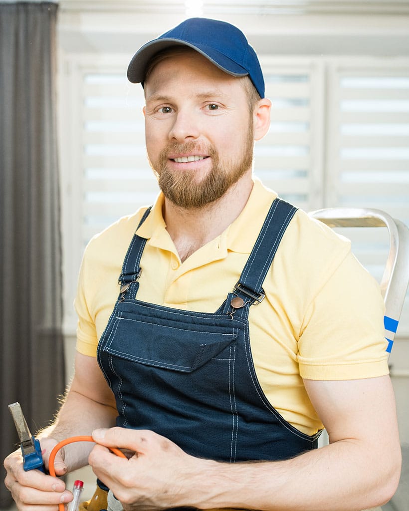 Smiling man in work overalls and cap holding tools.