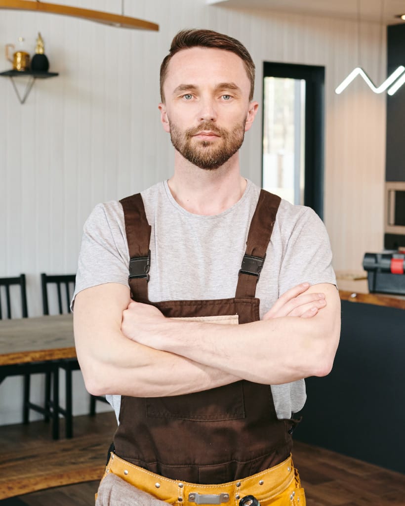 Man with beard wearing brown overalls and a grey t-shirt, arms crossed