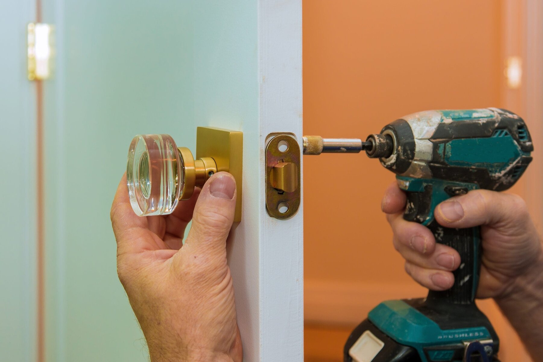Hands installing a clear glass doorknob and gold hardware with a power drill.