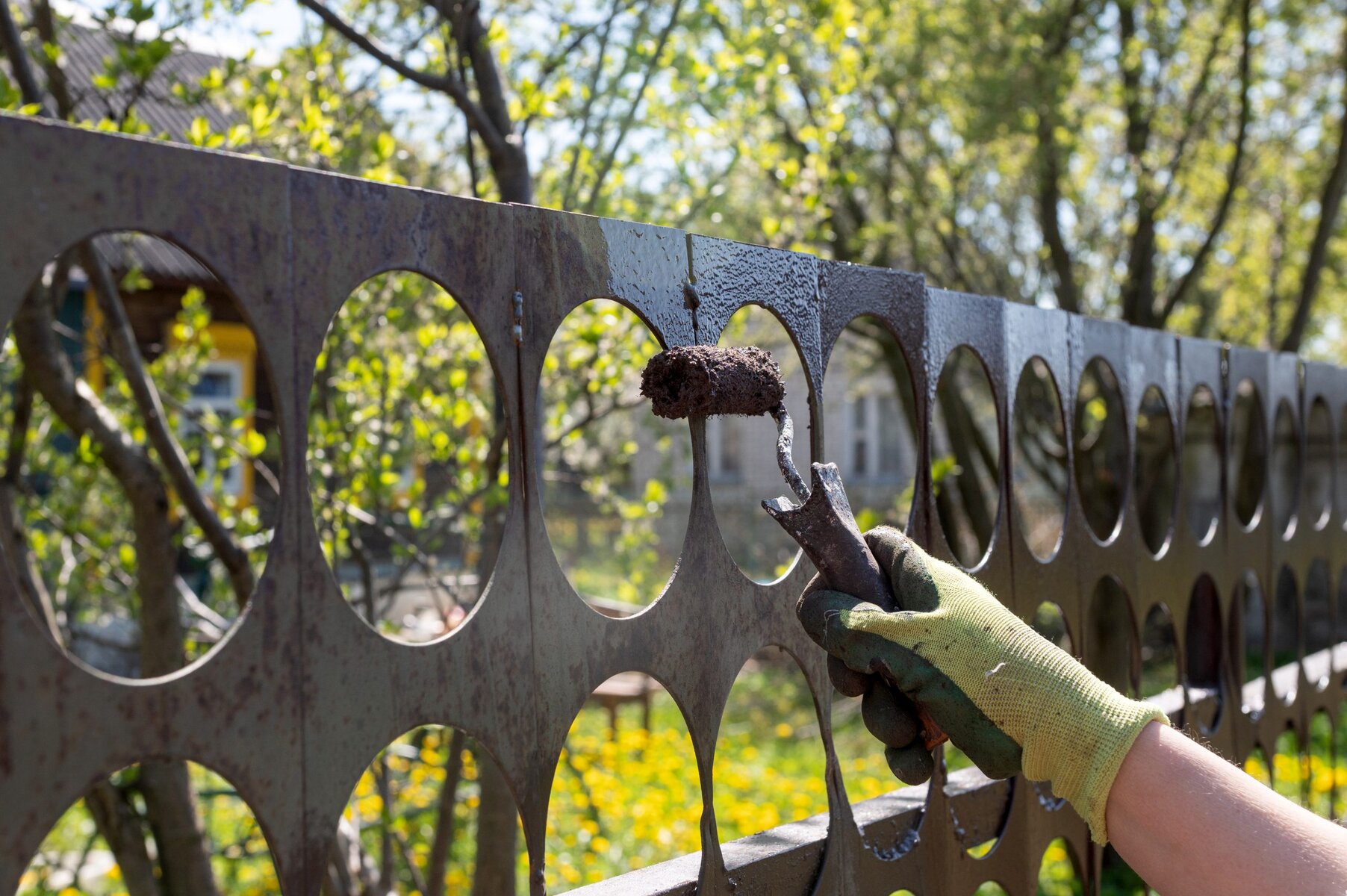 Hand in green glove painting a metal fence with a roller