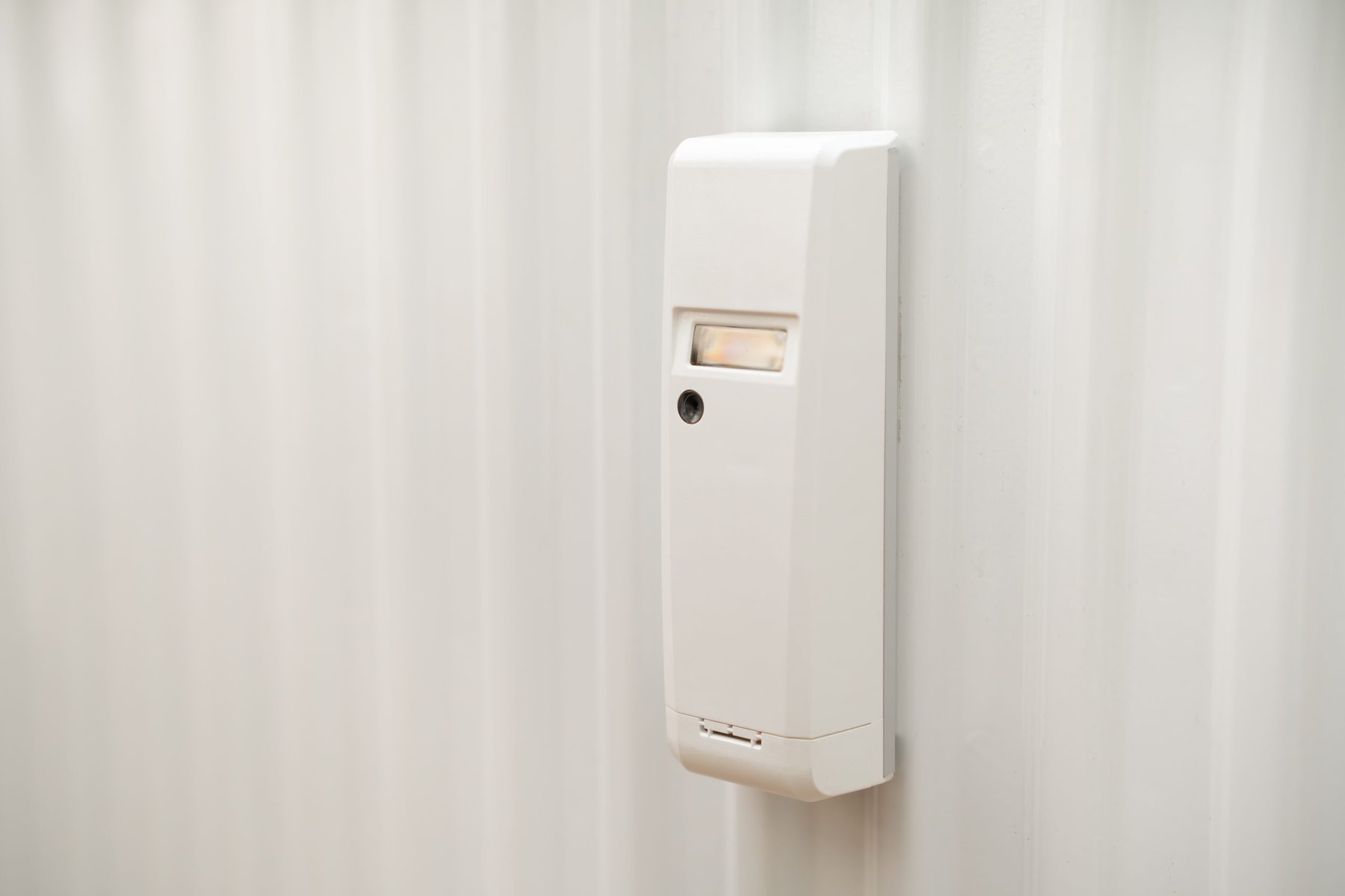 White rectangular security sensor with a small lens and light on a white wall.