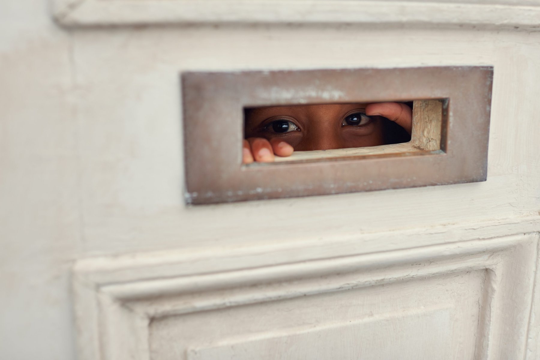 Child's eyes and fingers peeking through a letterbox on a white door.