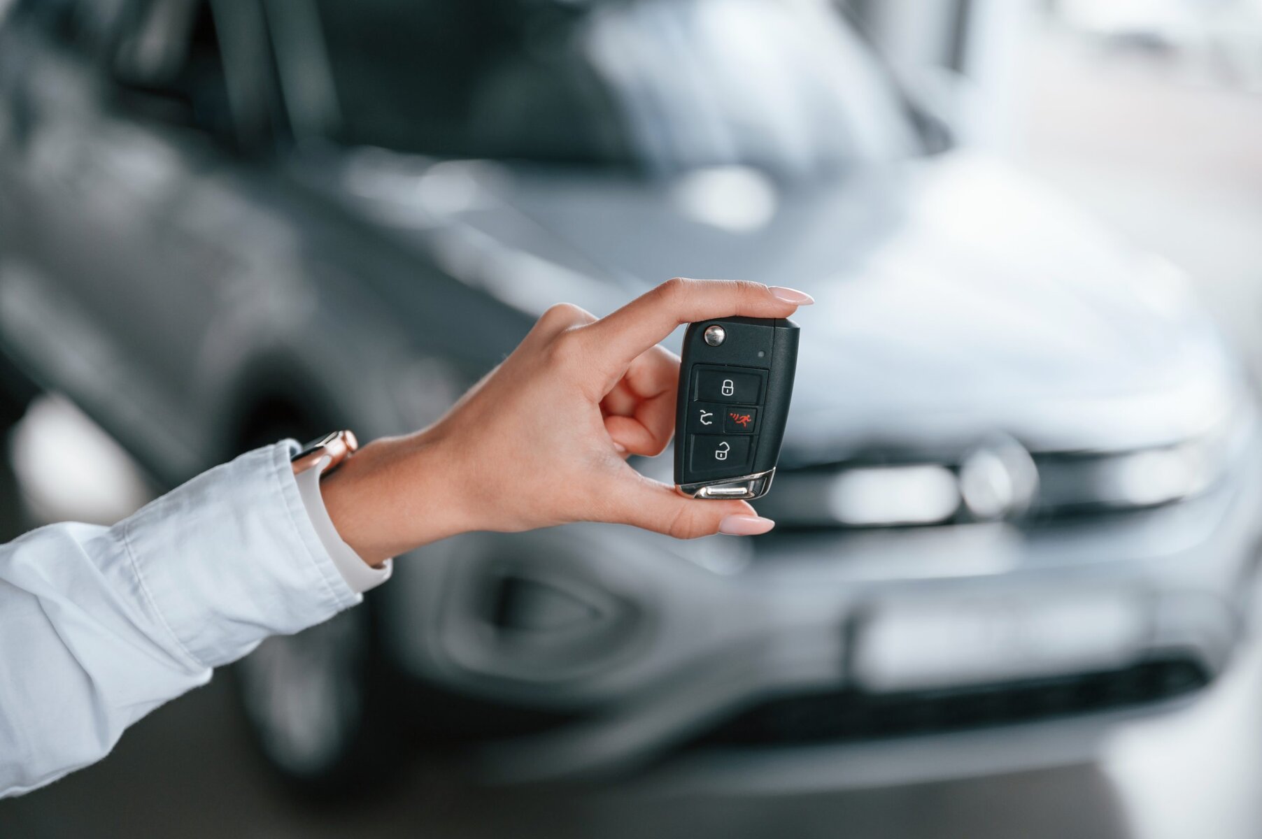 Hand holding a car key fob in front of a blurred car.