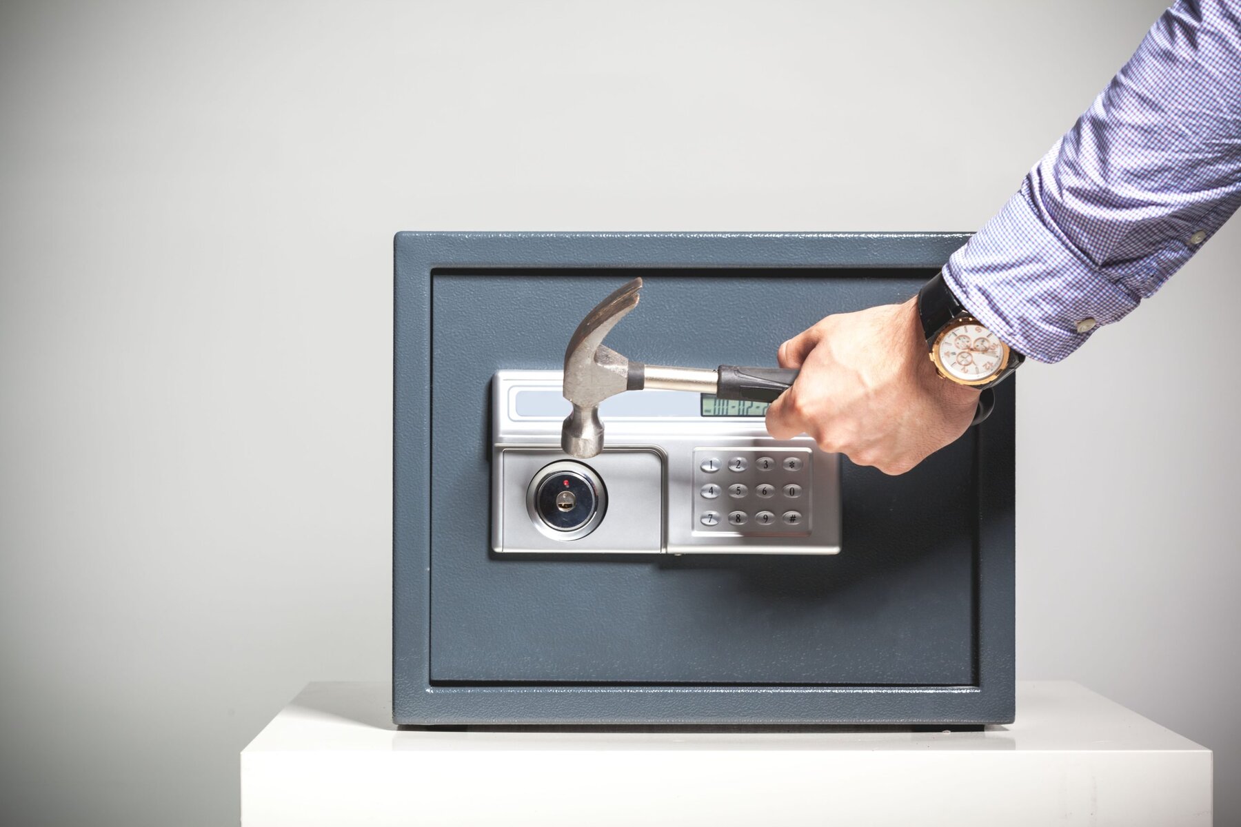 Man's hand with hammer attempting to open a grey safe with a digital keypad.