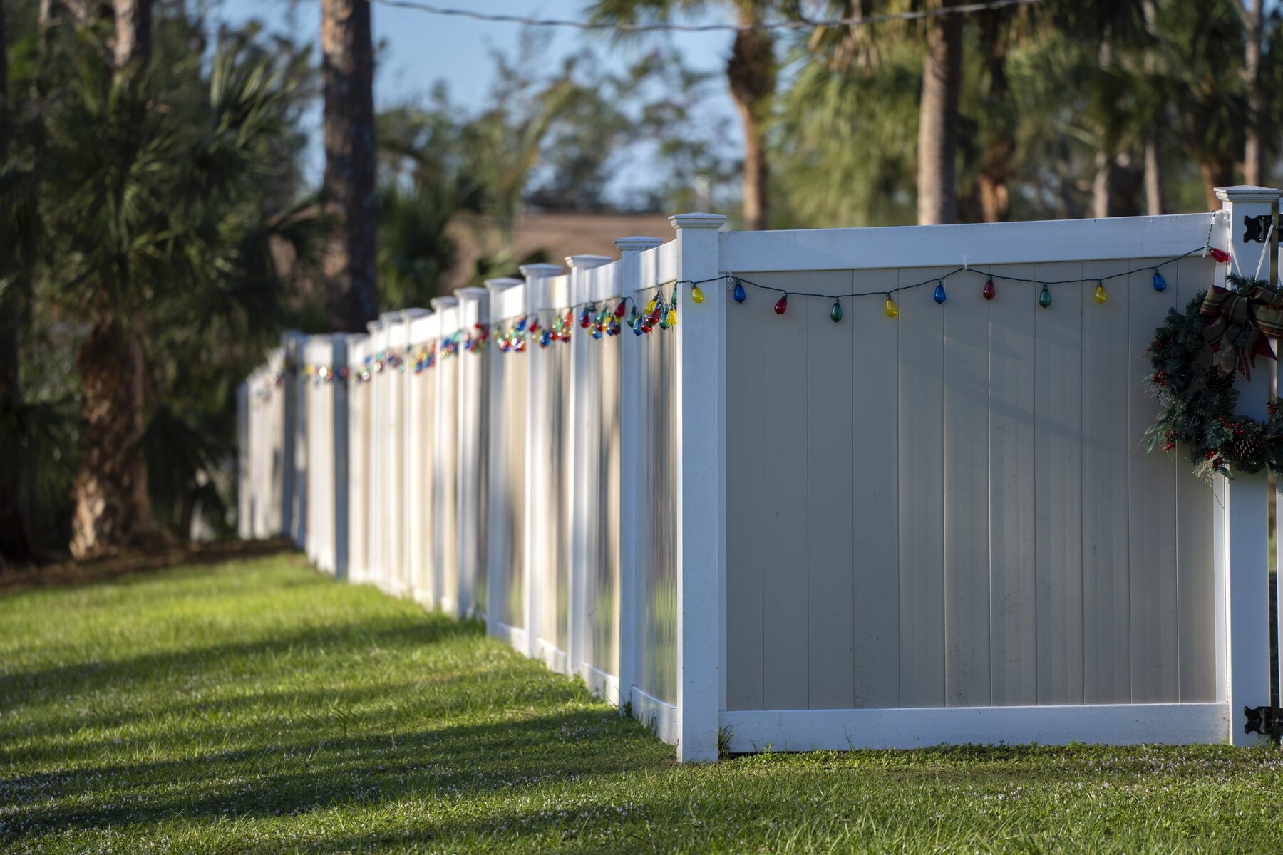 White fence with colorful string lights and a wreath on green grass.
