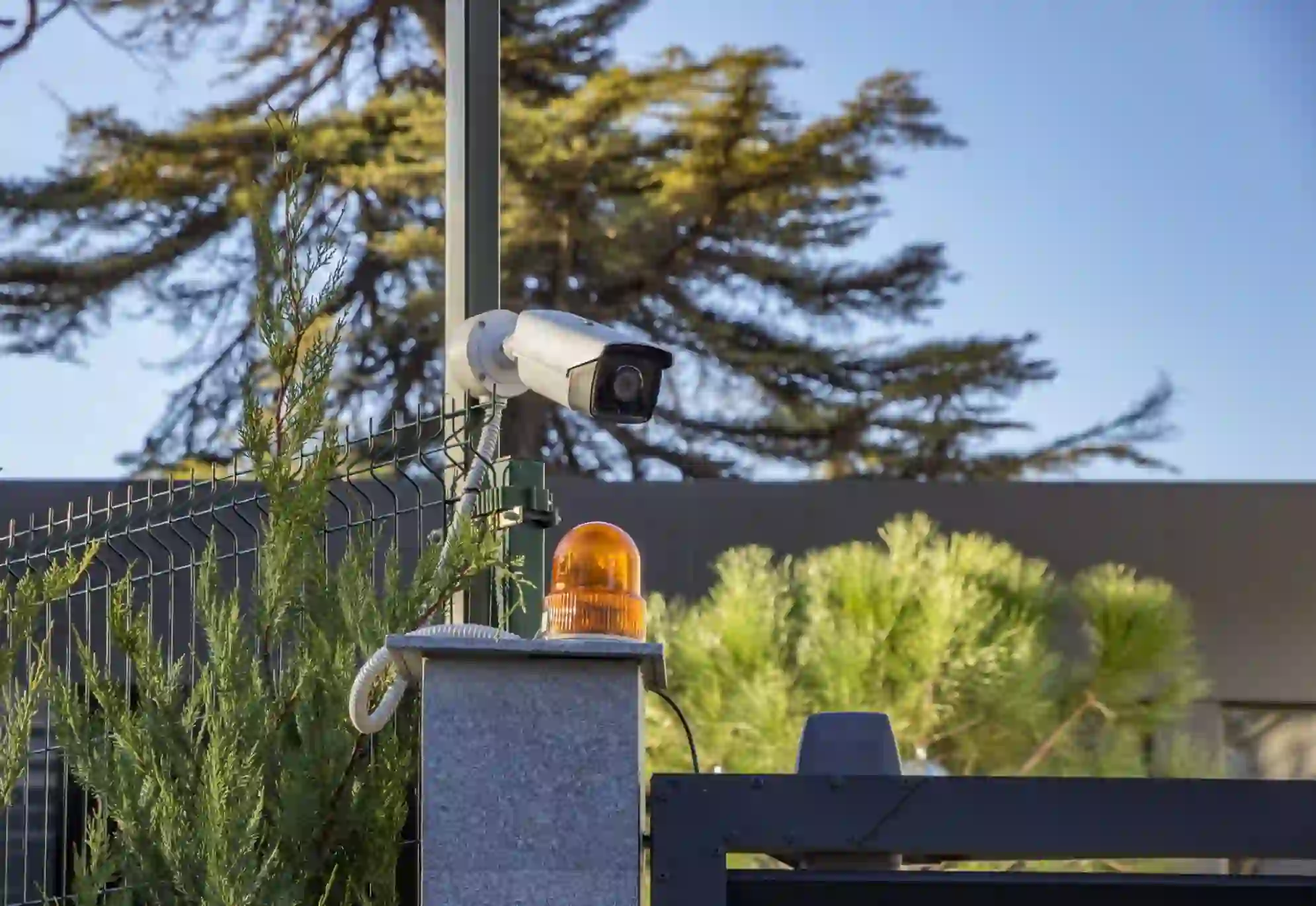 Security camera and orange warning light on a fence post.