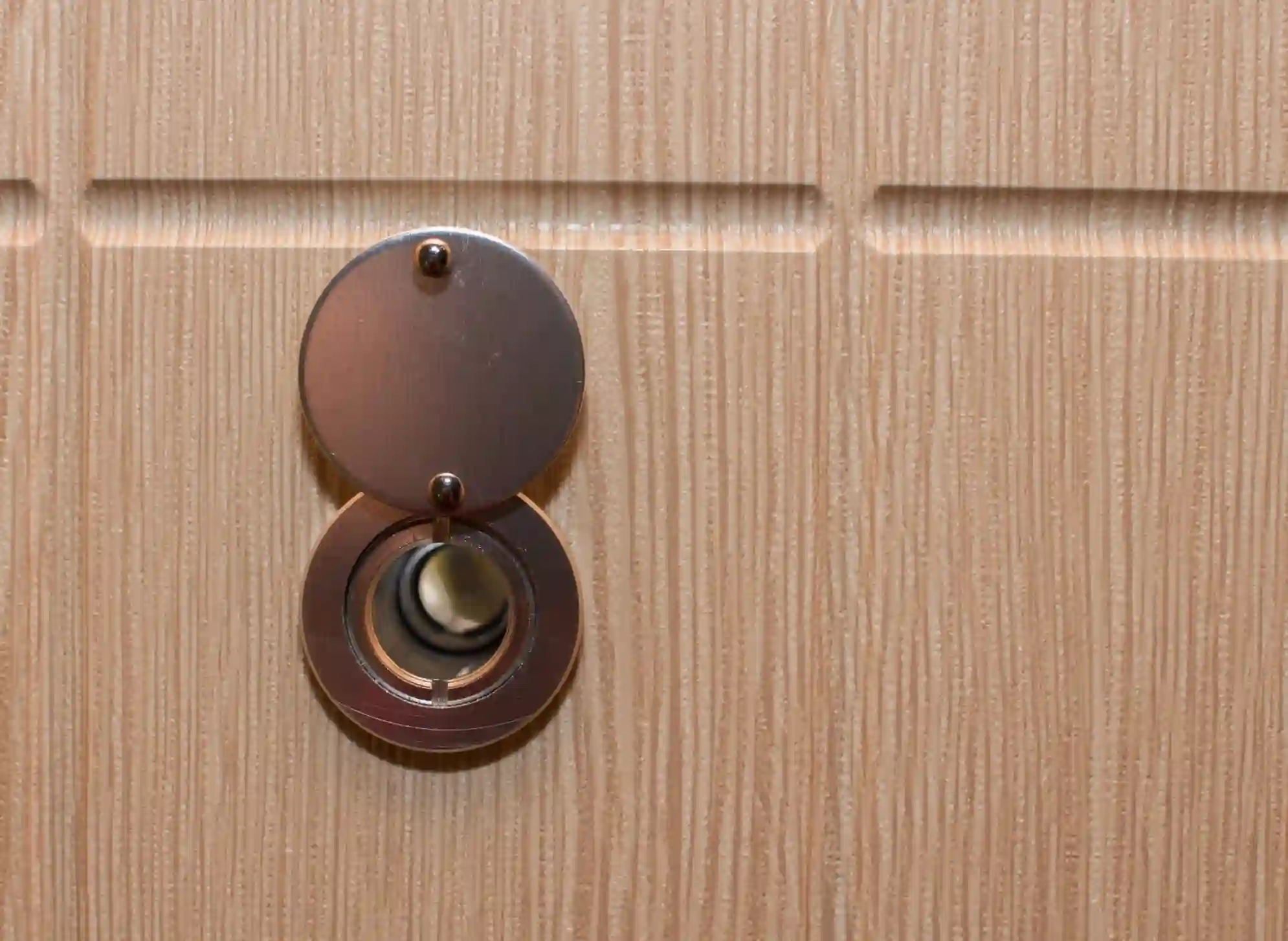 Close-up of a silver peephole on a light brown wooden door.