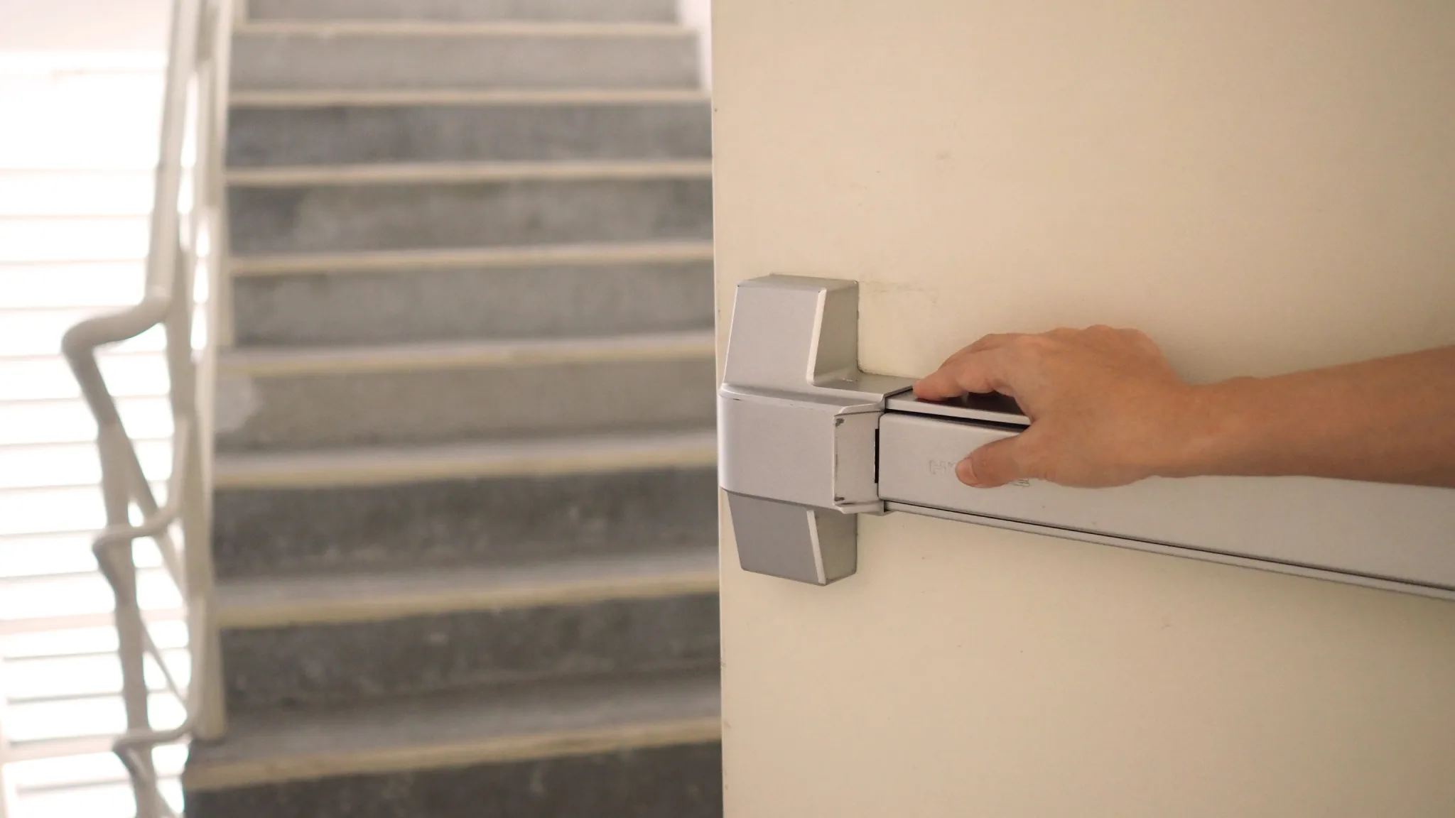 Hand pushing a silver panic bar on a door next to a concrete staircase.