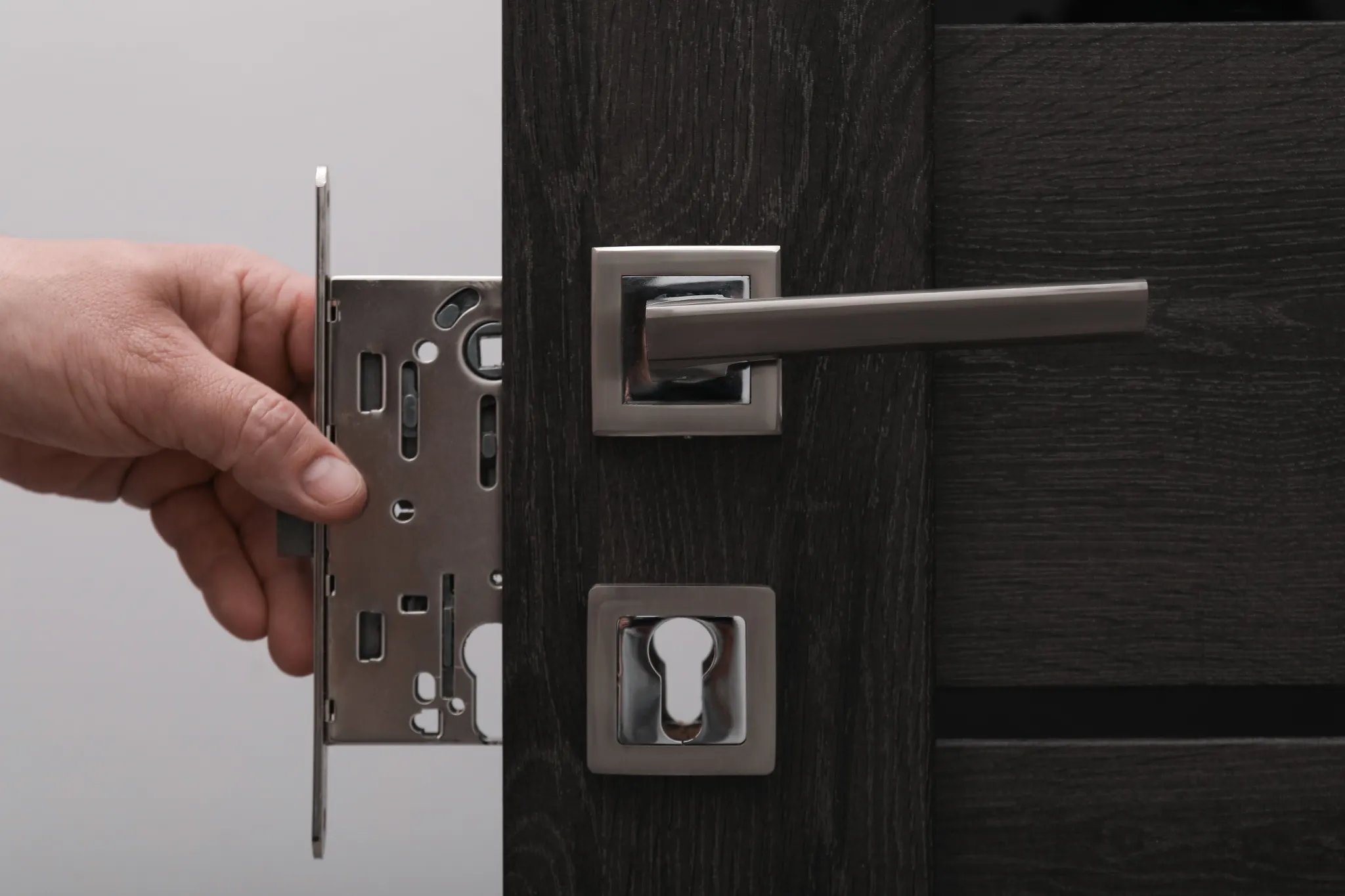 Hand holding a door lock mechanism next to a dark wooden door