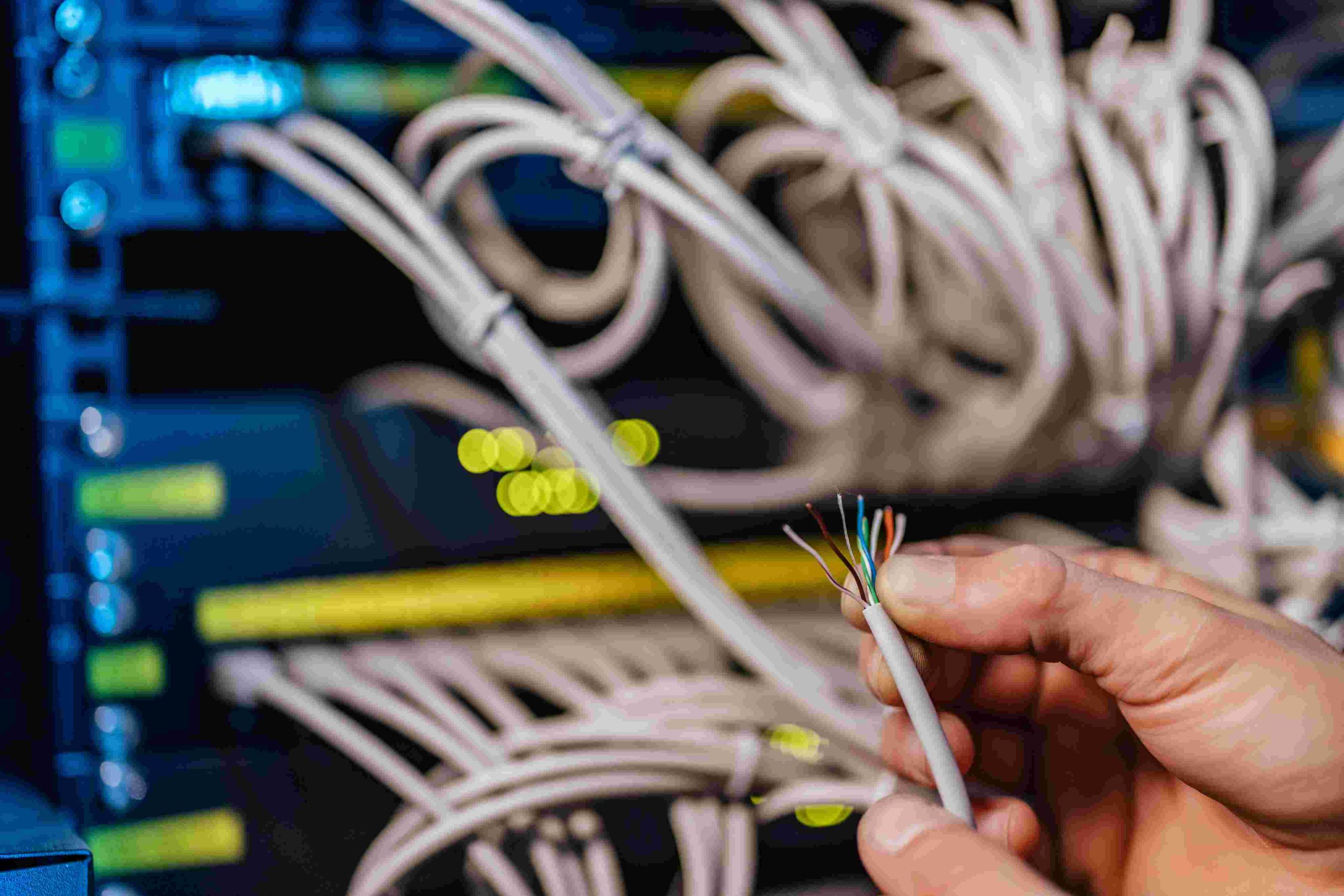Hand holding a cut network cable with colorful wires, server rack in background.