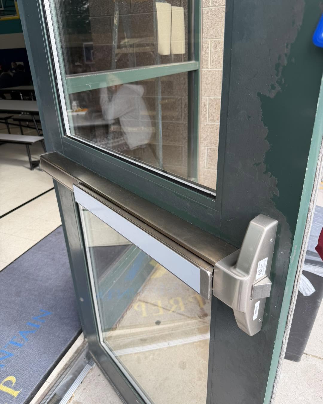 Green door with a push bar and window, showing a person inside.